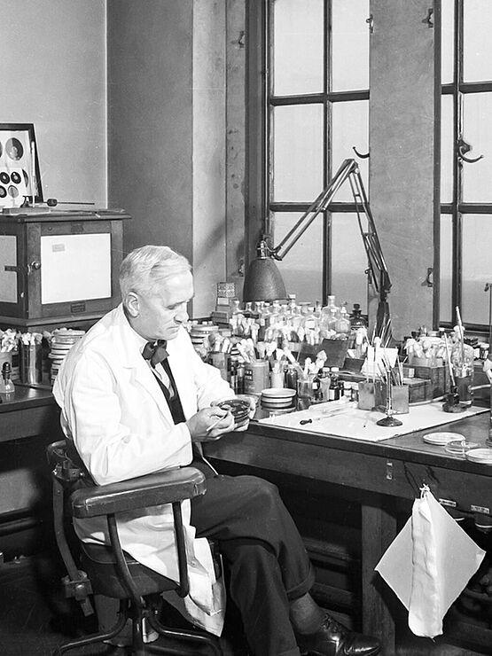 A scientist in a laboratory, examining a petri dish on his desk.