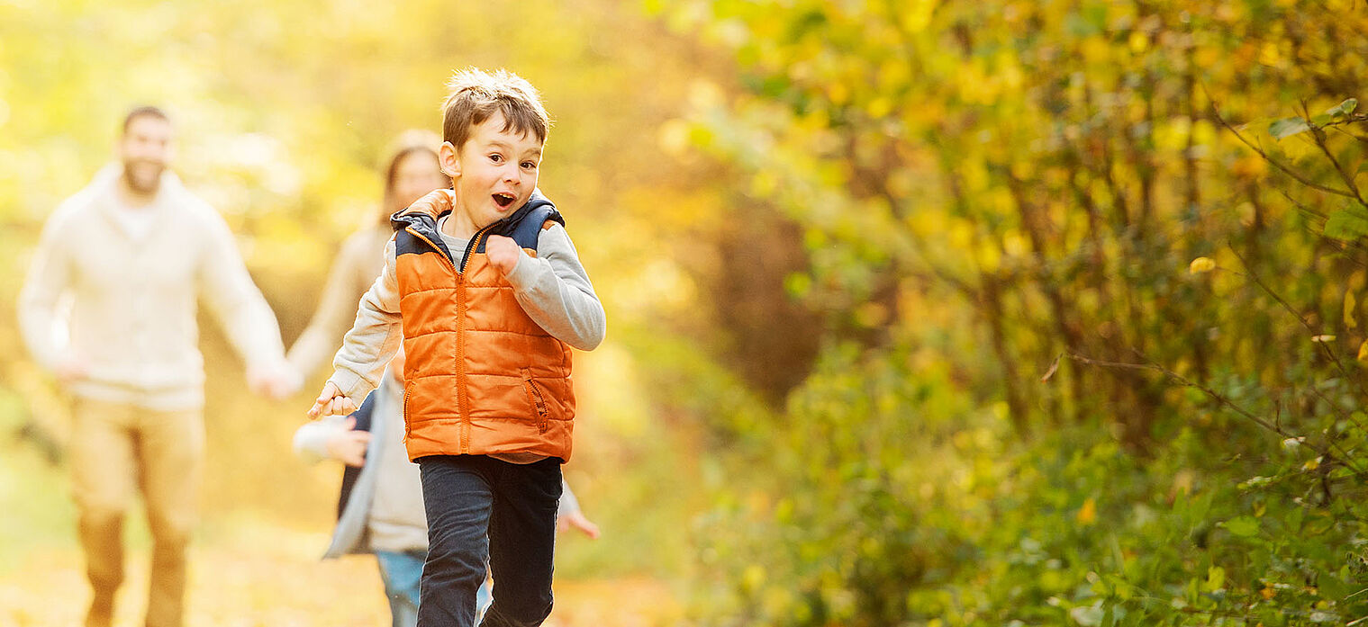 Young boy in an orange vest running joyfully on an autumn forest path, followed by a girl and an adult