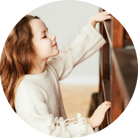 A young girl with long brown hair is reaching up to touch a shelf. She is wearing a cream-colored sweater with decorative bows on the sleeves