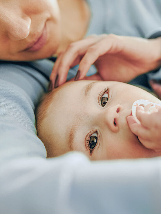 Close-up of a baby lying down, holding a pacifier, with a parent gently touching the baby's head