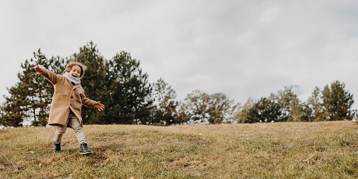 Child wearing a coat and scarf, running joyfully on a grassy hill with trees in the background on an overcast day