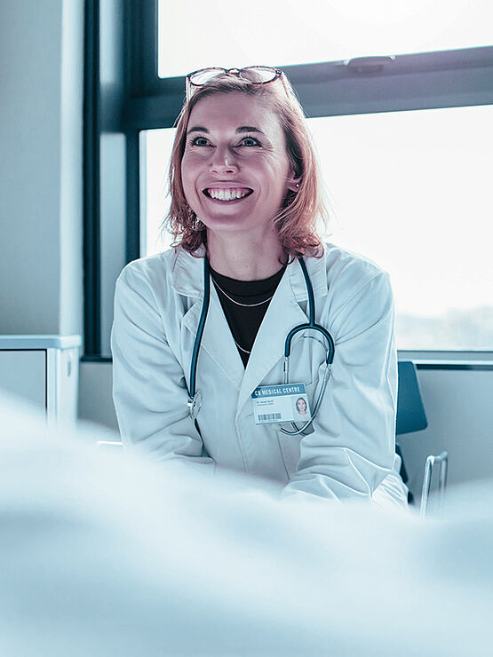 A smiling doctor in a white lab coat with a stethoscope, sitting and talking to a patient.