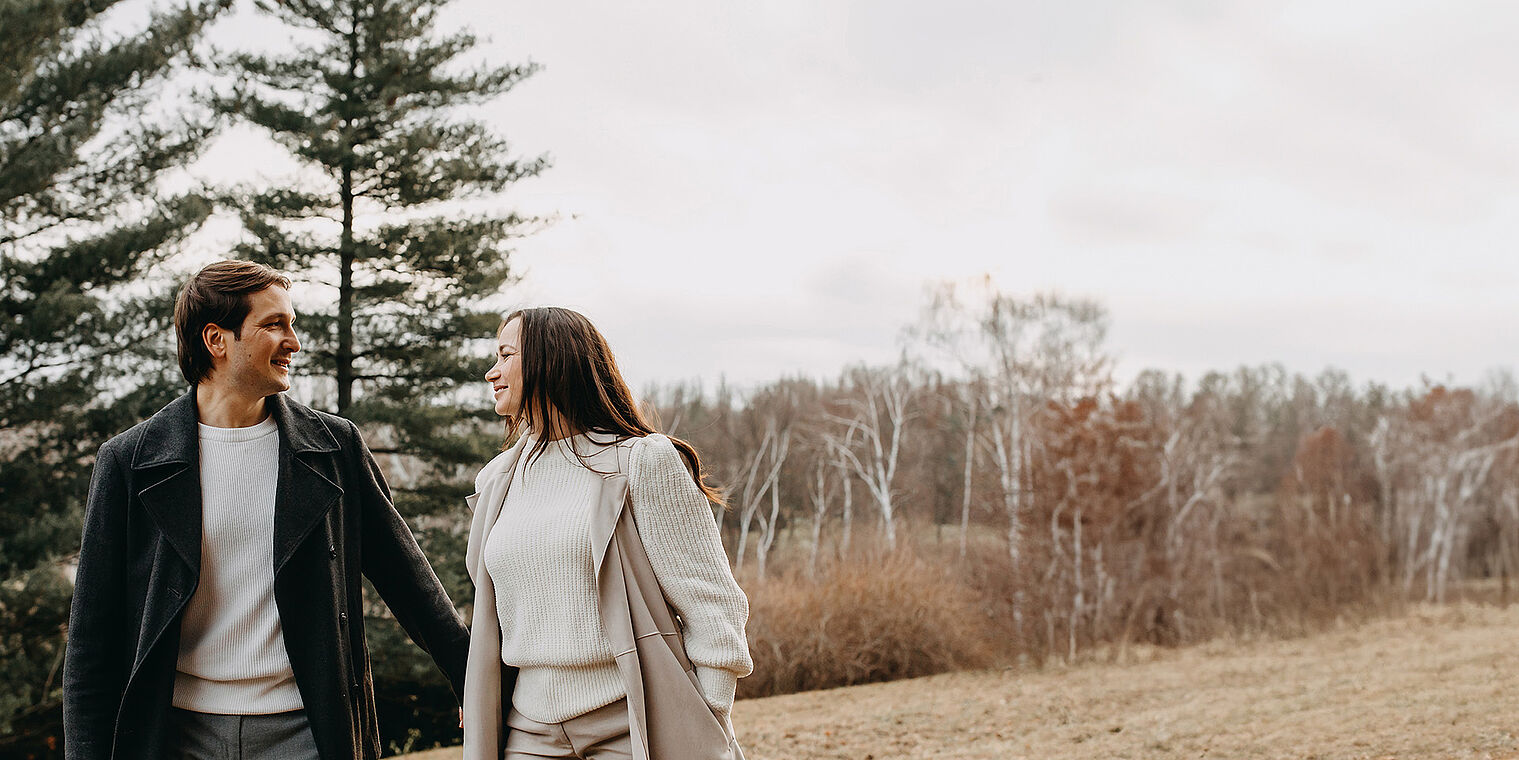 "A young couple dressed warmly in coats, walking and smiling at each other in an open field with trees in the background