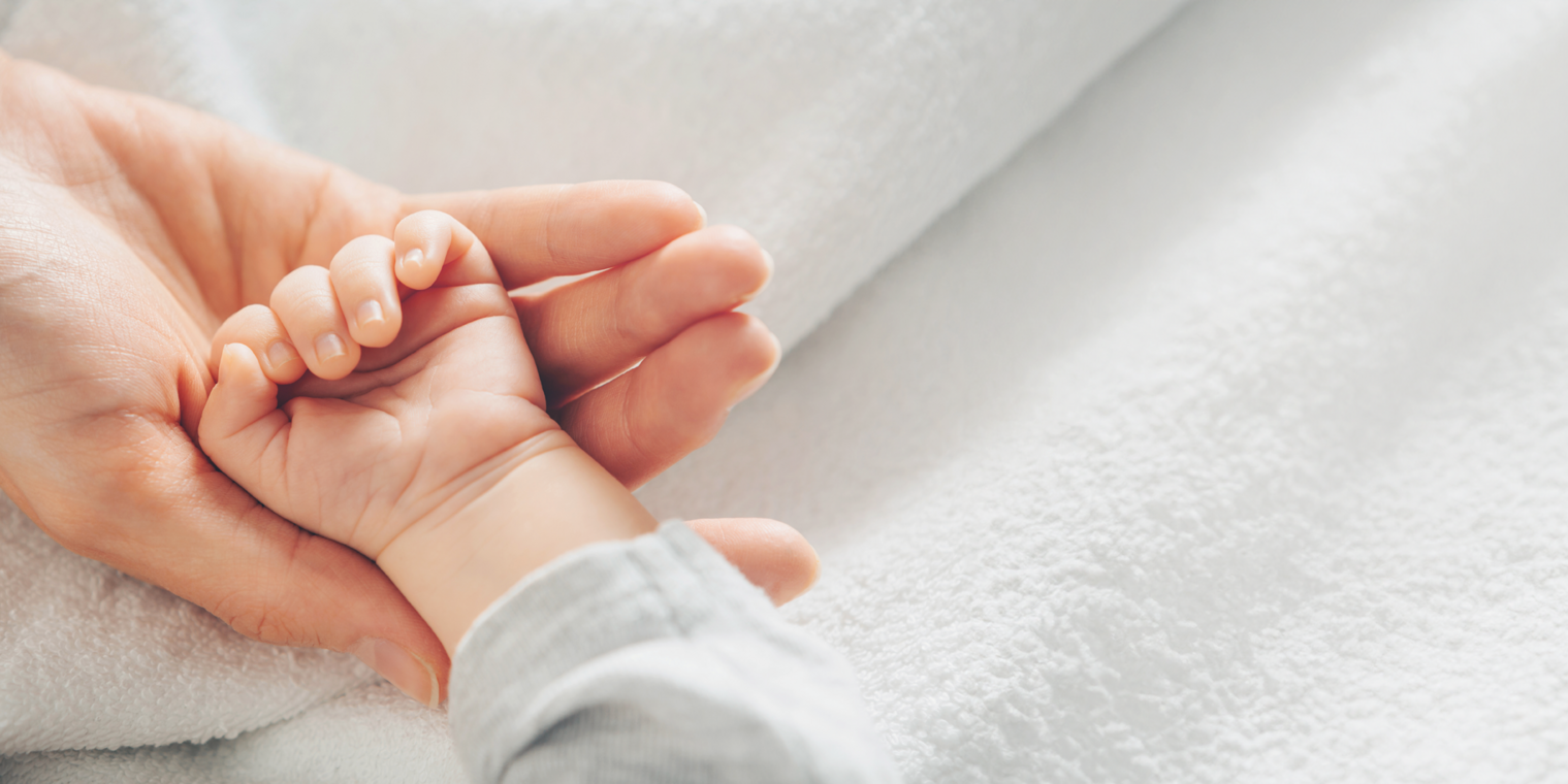 Close-up of a baby’s hand holding an adult’s finger, resting on a soft white blanket.