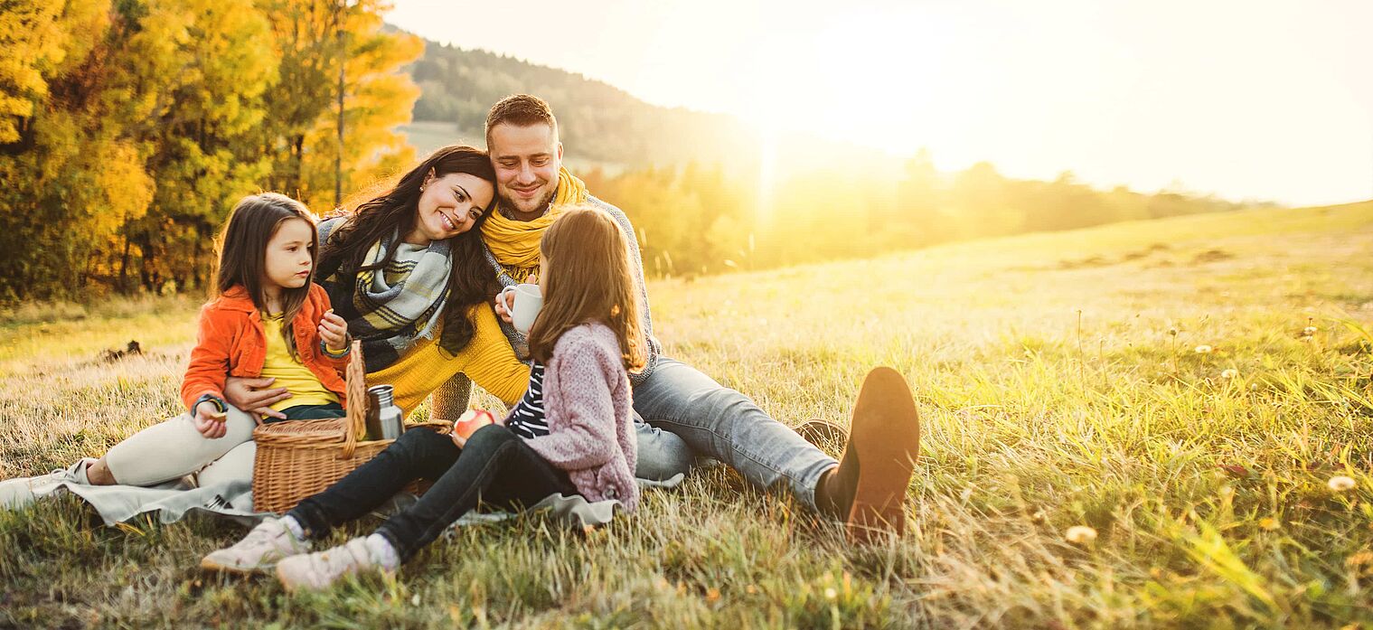 Family of four enjoying a picnic on a sunny autumn day, sitting on a blanket in a field with colorful fall trees in the background.